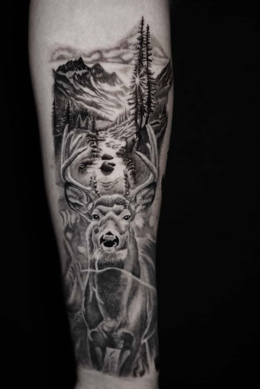 Black and white tattoo of a buck with antlers, mountains, river, and trees on an arm.