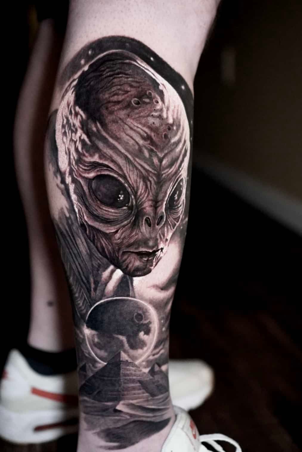 Alien tattoo on a leg, with a detailed gray alien face and pyramid scene below.