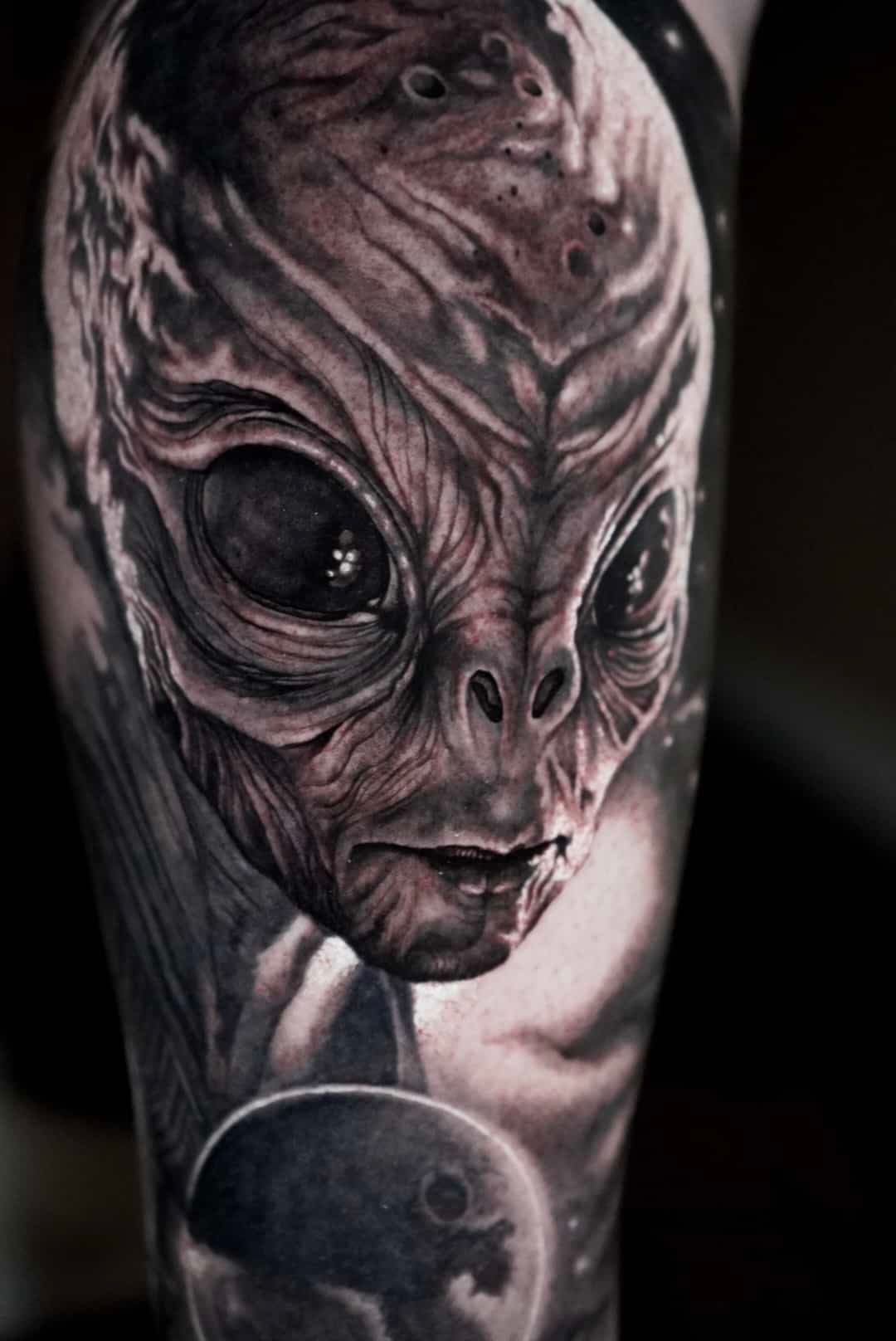 Black and gray alien face tattoo with large dark eyes and a planetary background.