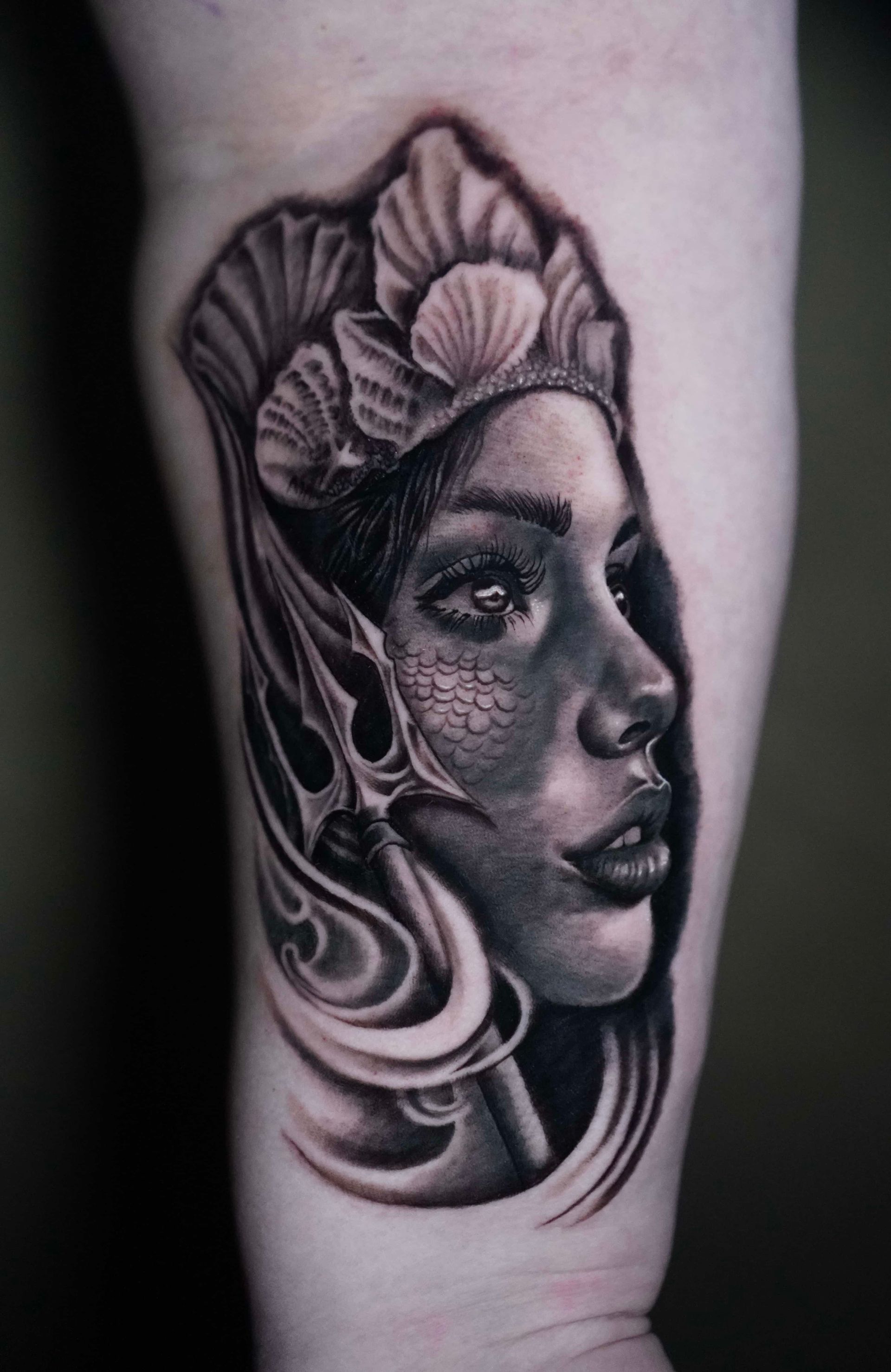Mermaid tattoo on arm in black and gray, wearing seashell crown and holding trident.