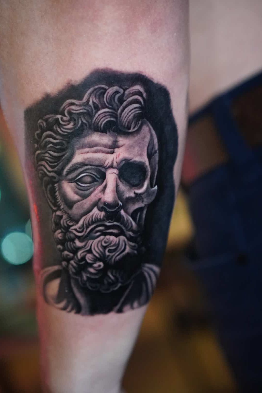 Tattoo of a stone bust with one eye and part of the skull exposed, in grayscale on arm.