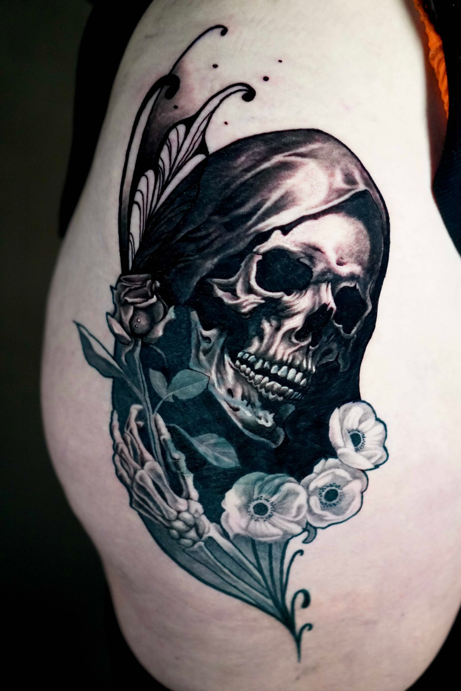 Skull tattoo with flowers, dark shading, and skeletal hand on a person's thigh.