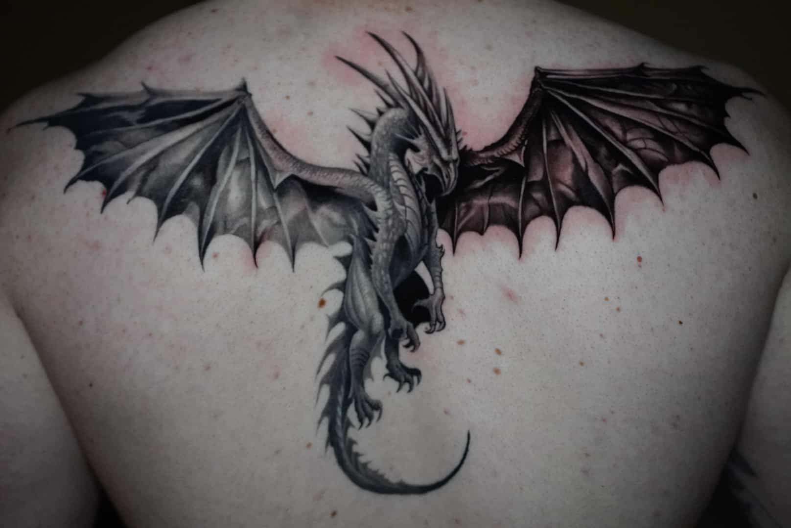 Dragon tattoo with outstretched wings on a person's back; black, grey, and white shading.