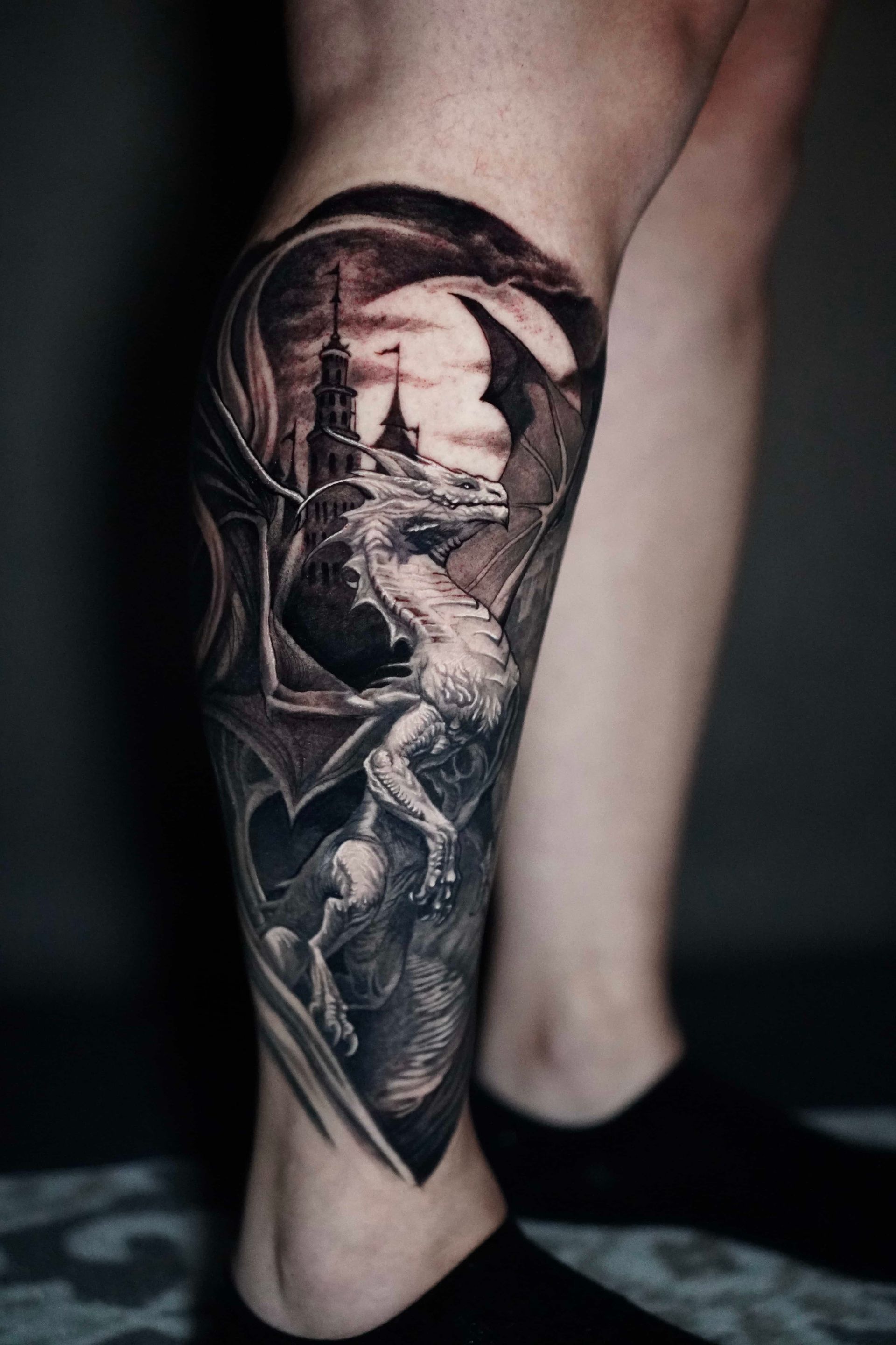 Detailed black and grey dragon tattoo on a leg, with a castle under a moonlit sky.
