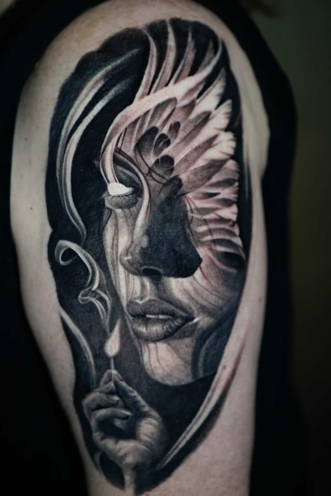 Black and white tattoo of a woman's face with a wing-like design, holding a lit match; on a person's upper arm.