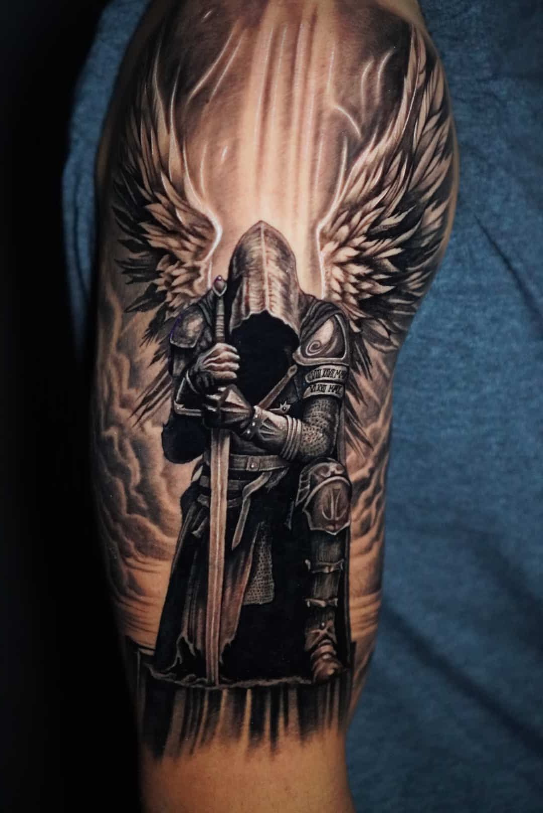 Tattoo of armored angel kneeling, holding sword, with large wings, in a cloudy setting, grayscale.