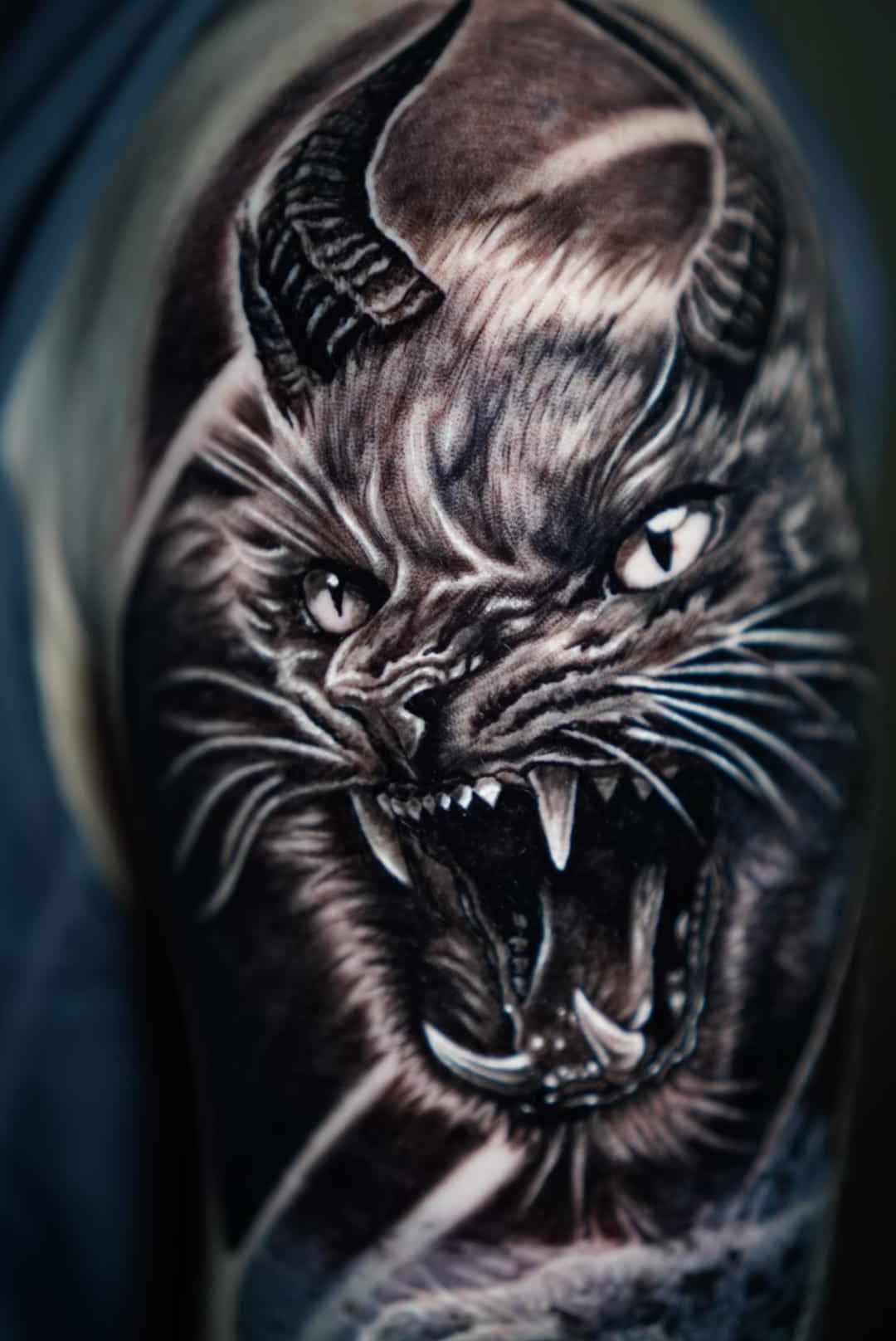 Black and gray tattoo of an angry feline creature with horns, bared fangs, and piercing eyes.