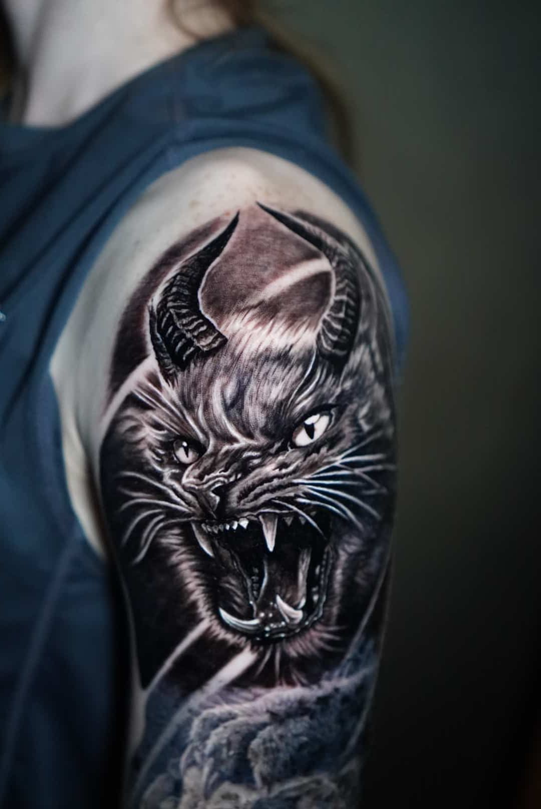 Black cat tattoo with horns, bared teeth, and intense eyes on a person's upper arm.