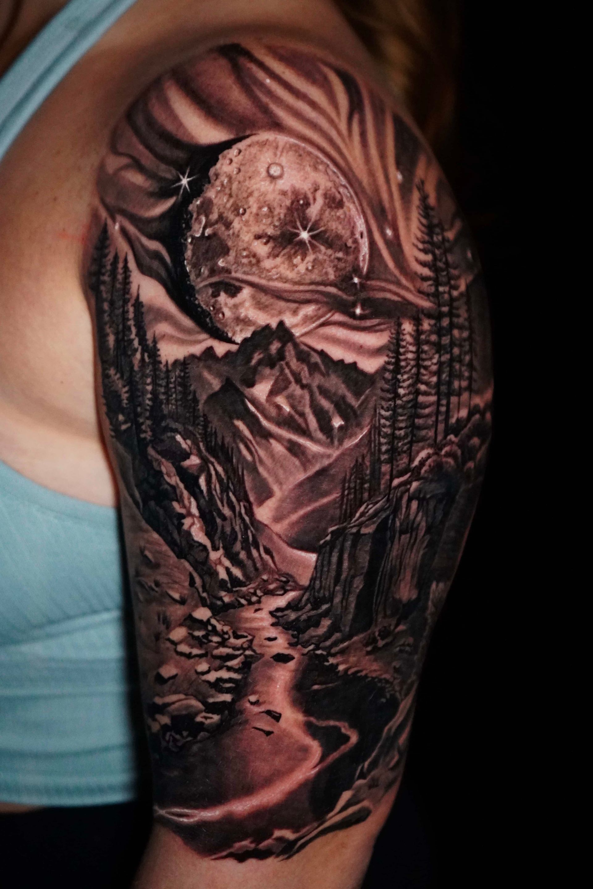 Detailed black and grey mountain landscape tattoo with moon, stars, and flowing river on a person's upper arm.