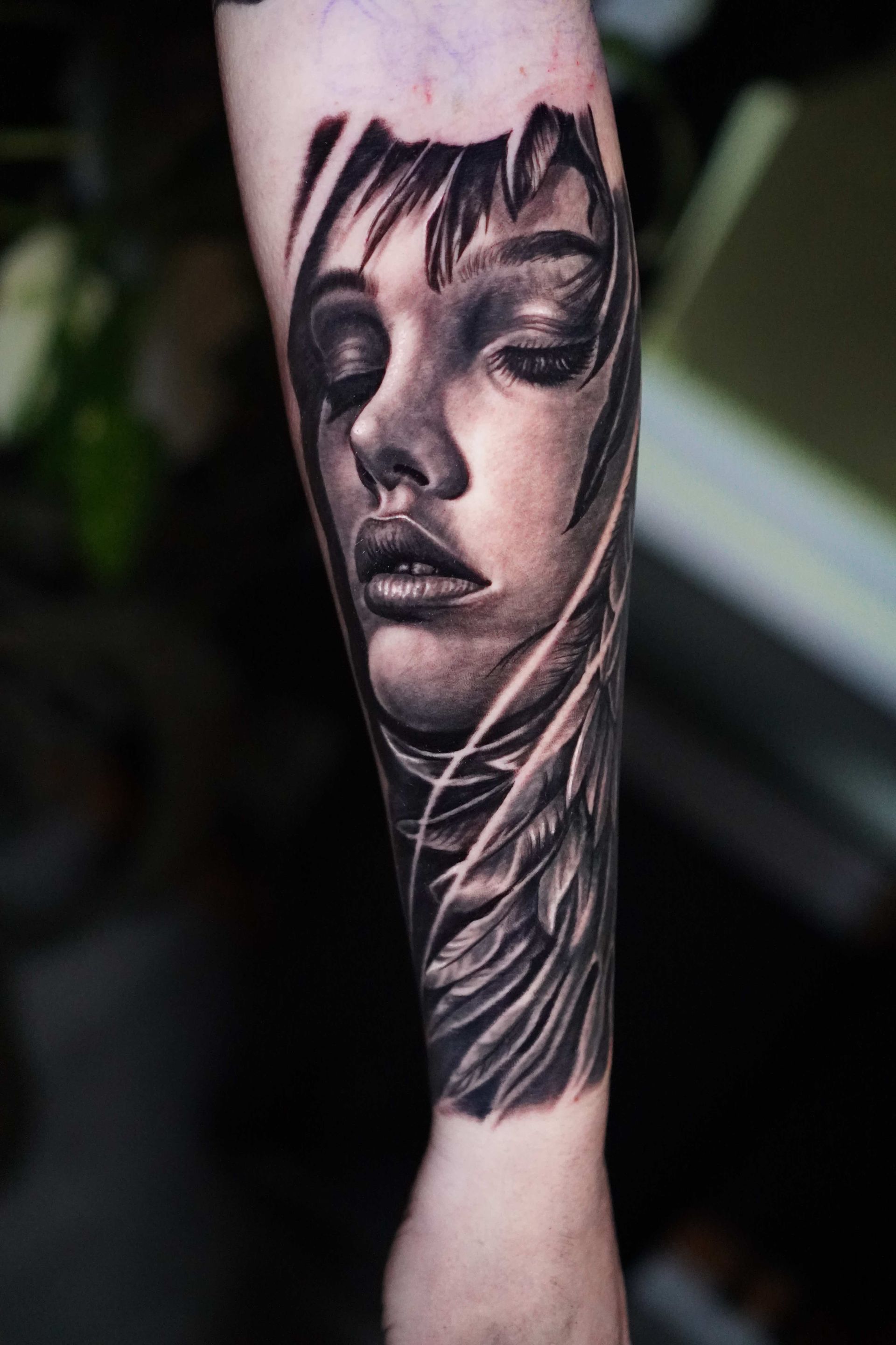 Black and white tattoo of a woman's face on arm, featuring closed eyes and flowing hair. Angel wings are also present.