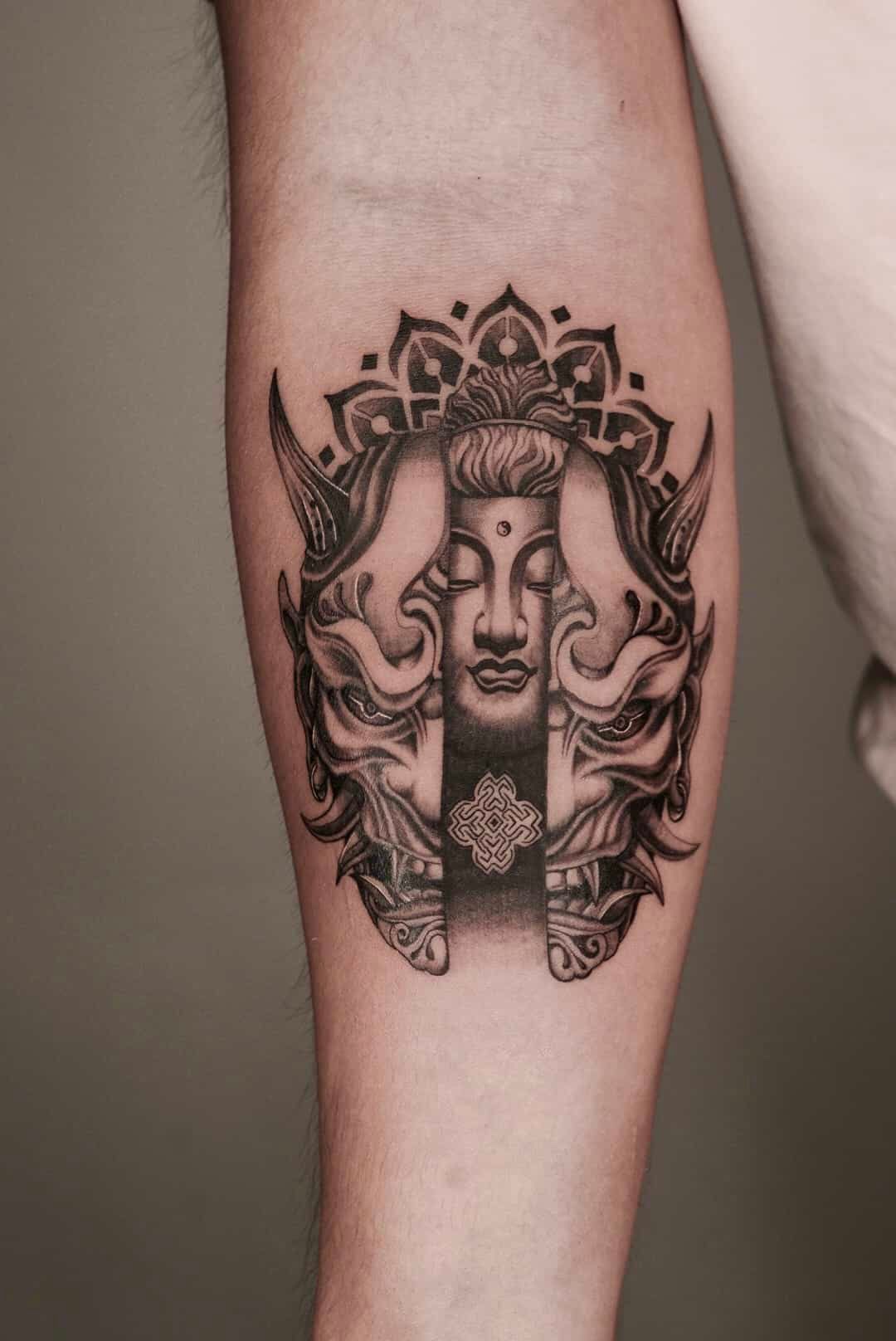 Tattoo of a Buddha face framed by ornate elements and a decorative top, on a forearm.