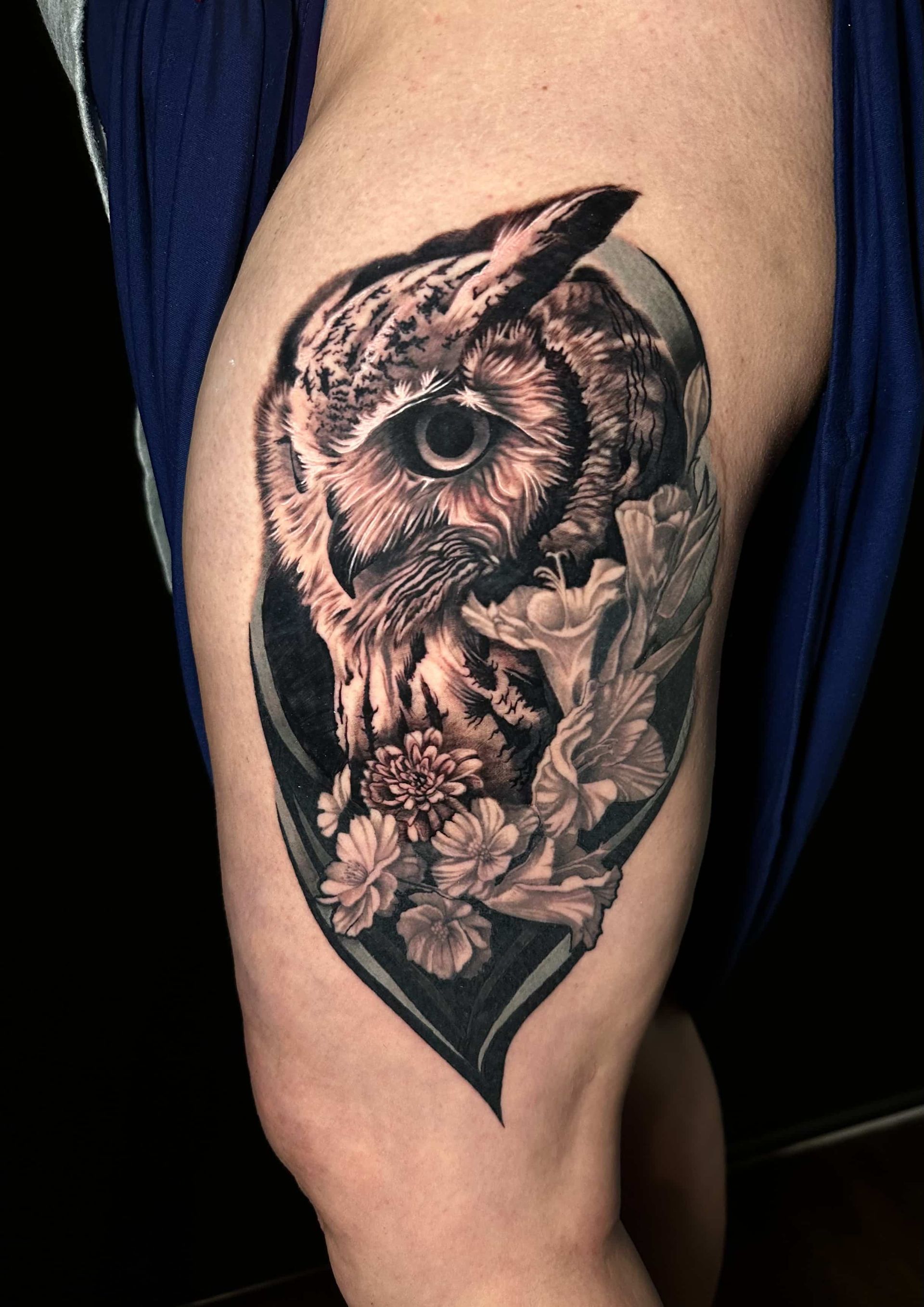 Realistic owl tattoo on a thigh, with flowers. Black and gray shading with a dark oval background.