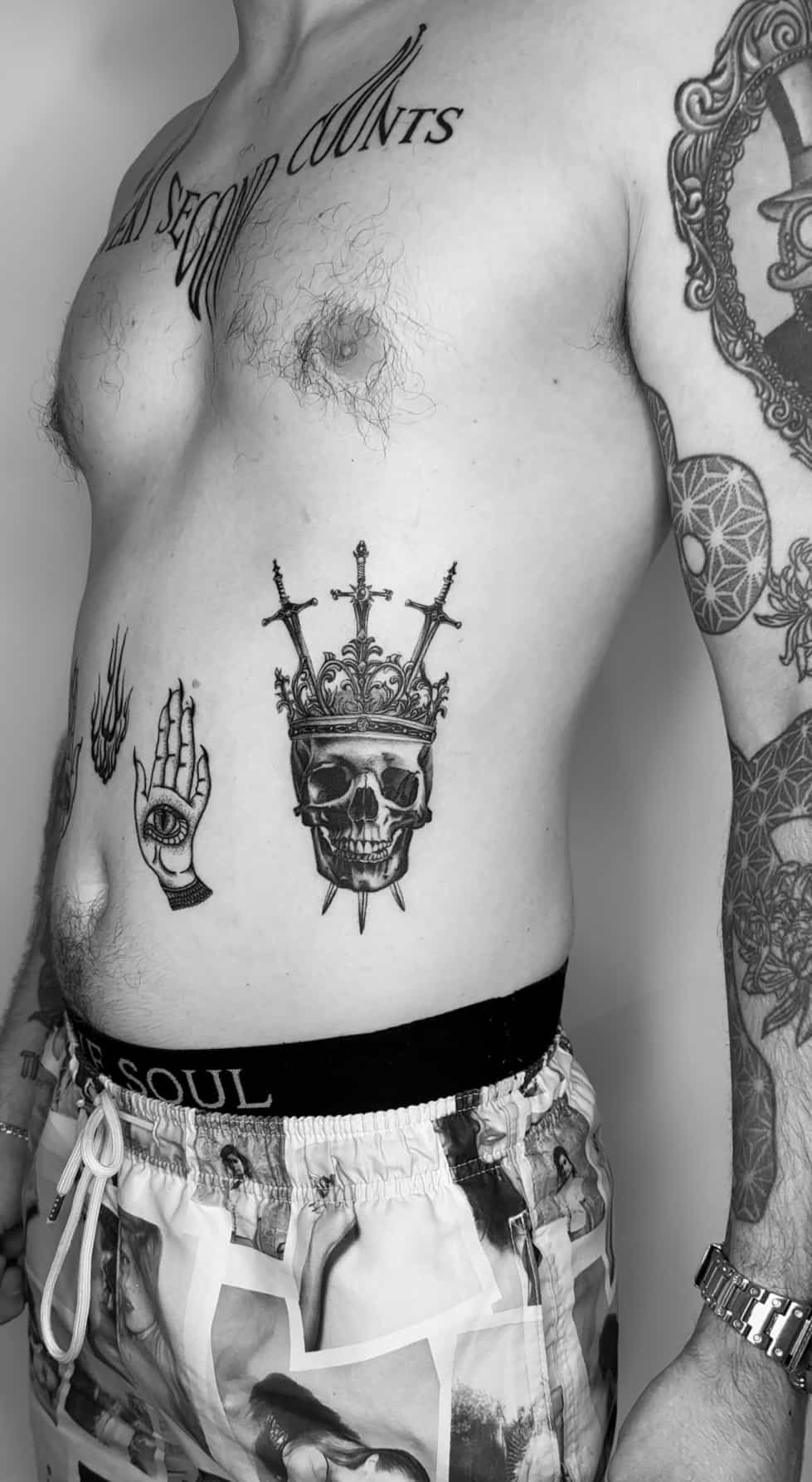Torso with tattoos including skull crown, hand, and chest script, wearing patterned shorts.