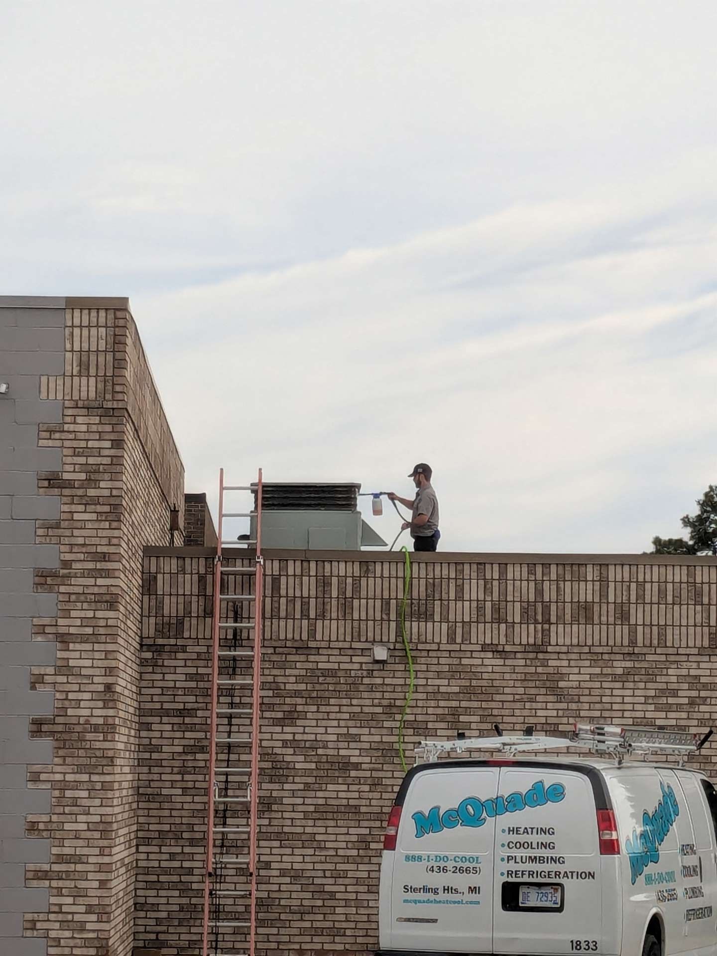 A person stands on a brick rooftop, inspecting something with equipment. A ladder leans against the building, and a white van with the 