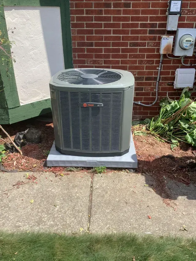 An air conditioner is sitting on the sidewalk in front of a brick building.