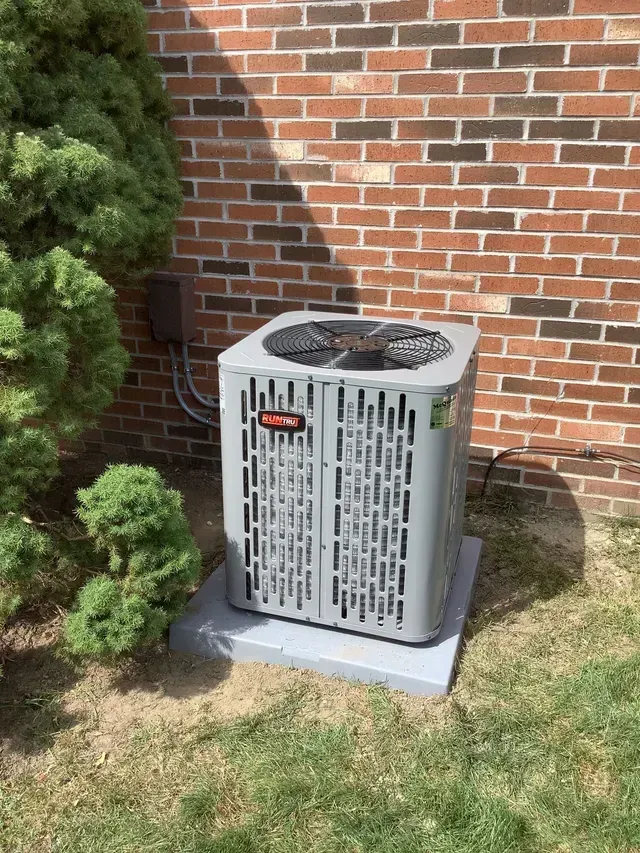 A gray air conditioner is sitting in front of a brick wall.