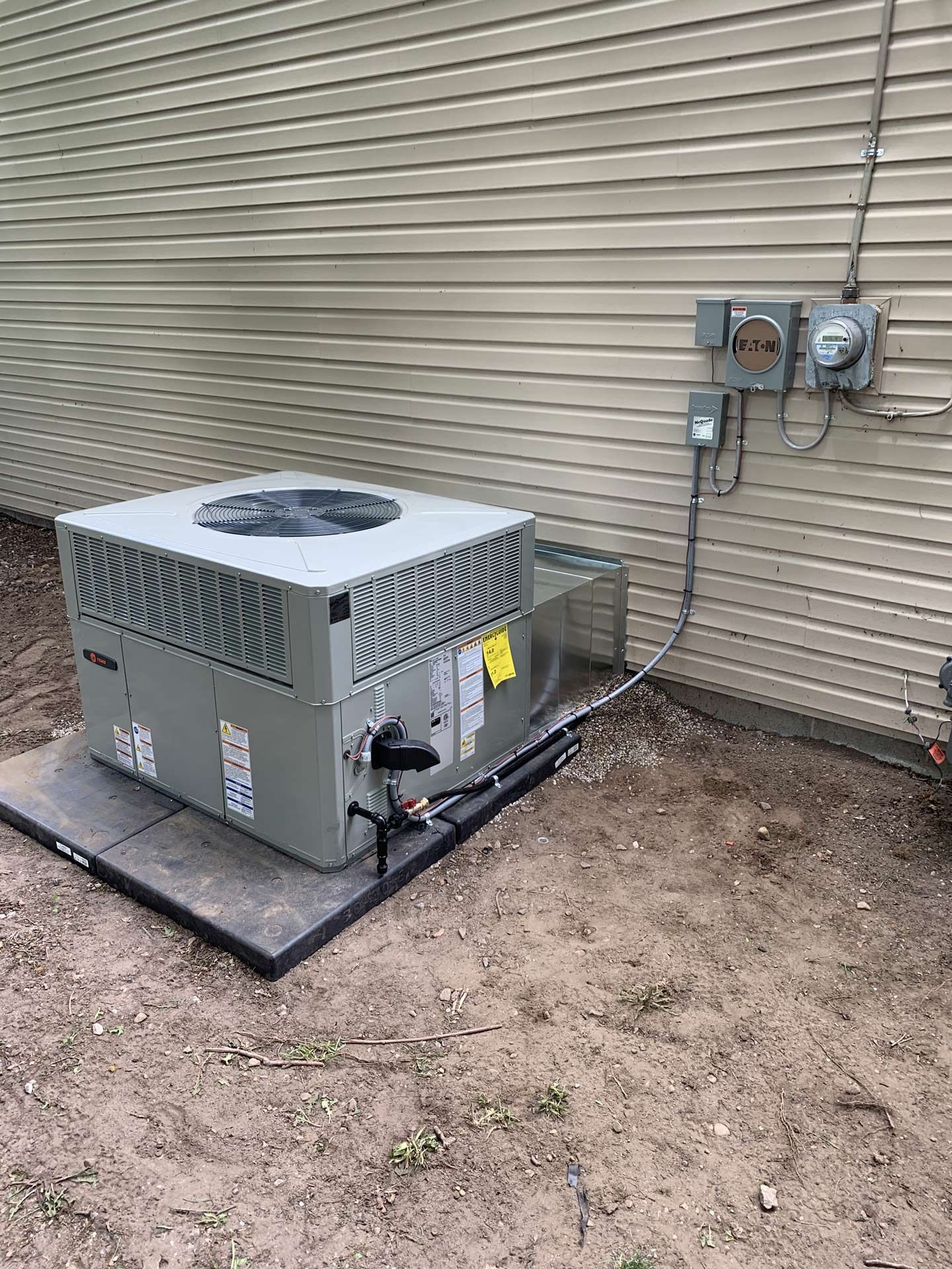 Outdoor air conditioning unit next to a building with electrical components on the wall. Unit is on a black pad in a dirt yard.