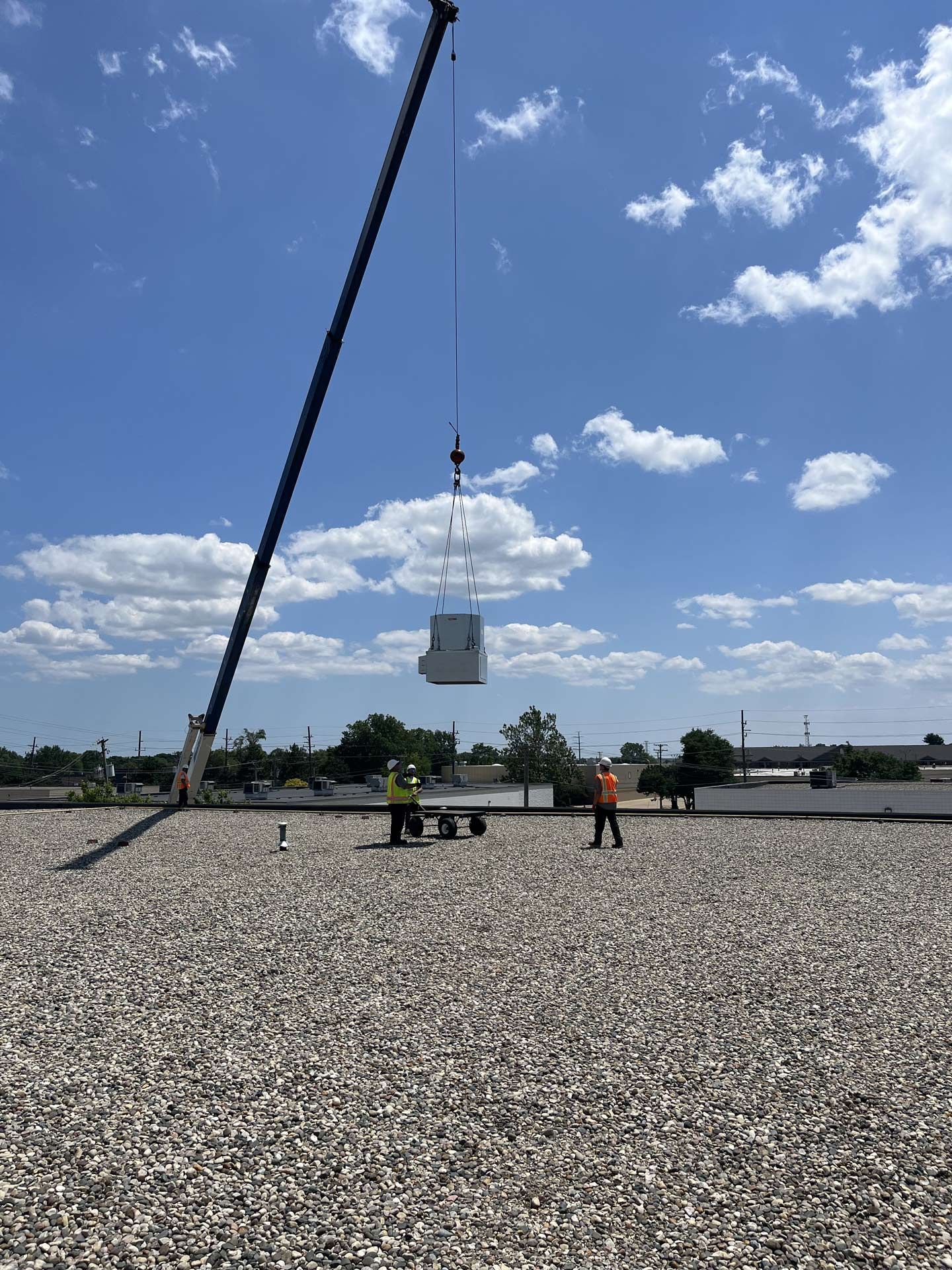 A crane lifting an HVAC unit onto a rooftop. Two workers in safety vests are on the roof.