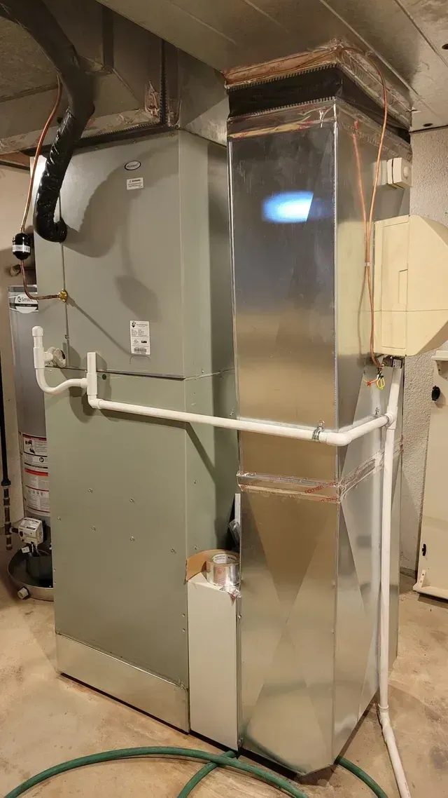 A large stainless steel heater is sitting in a basement next to a green hose.