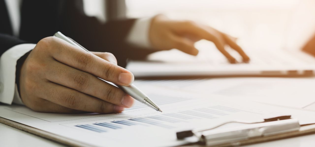 Person in a suit writing on documents with a pen, possibly reviewing financial data.