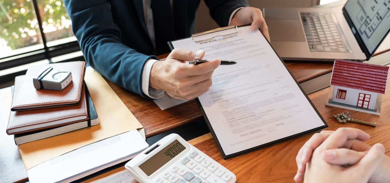 A person in a suit points to a document for a person with their hands clasped. A laptop, calculator, and model house are on the table.