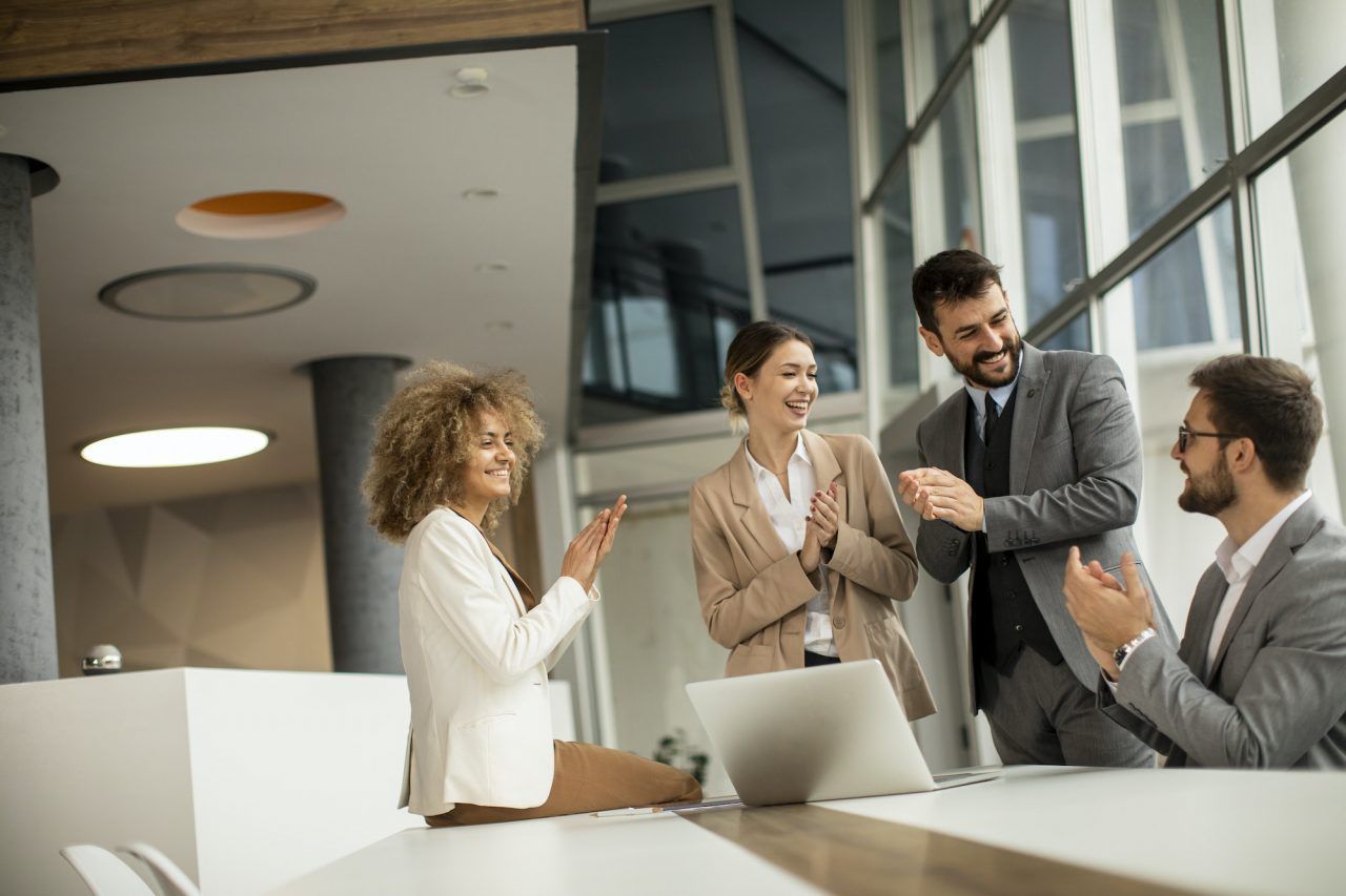 Four colleagues in suits celebrate a successful project, clapping and smiling in a modern office with large windows.