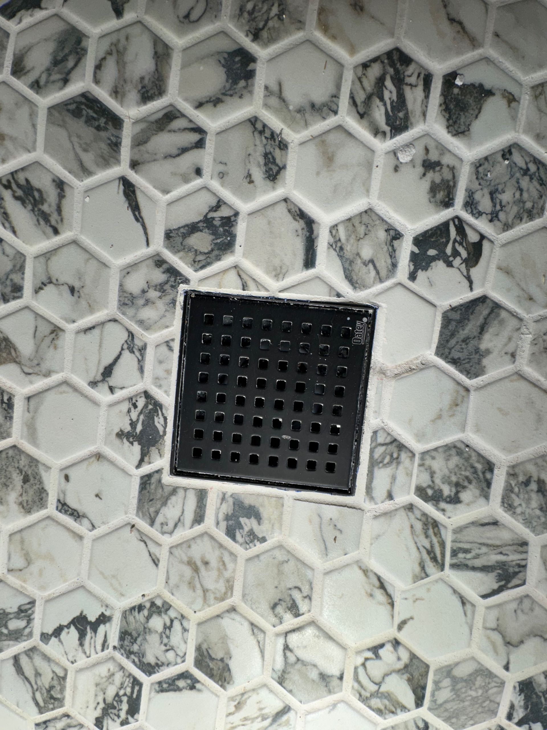 A close up of a shower floor with hexagon tiles and a black drain.
