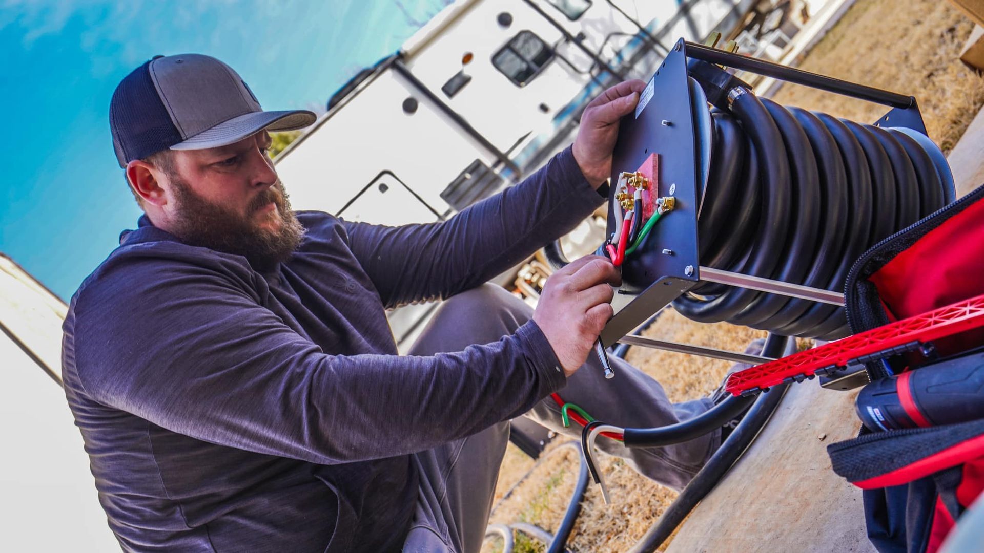 Man with beard working on a black electrical cord reel, near a camper.