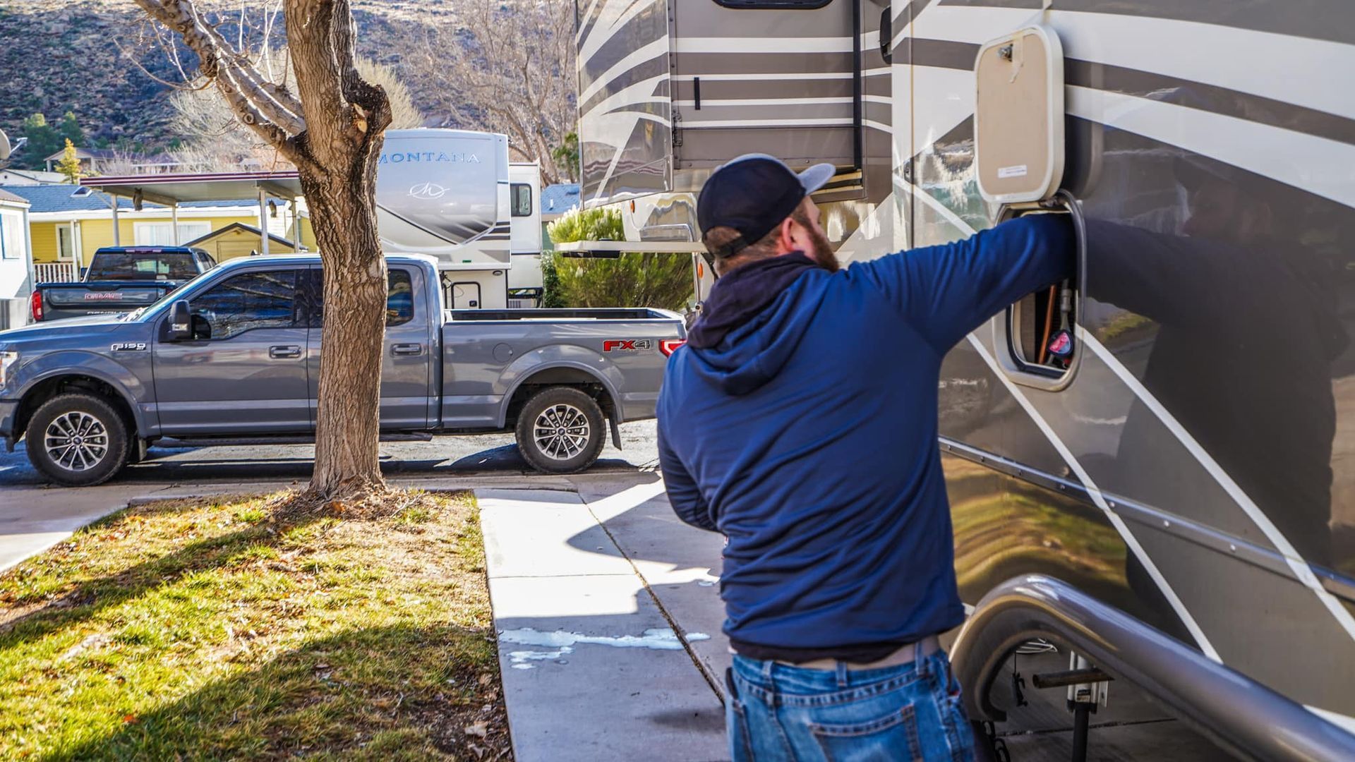 Man in blue shirt, jeans by RV, adjusting something. Grey truck parked in the background, daytime.