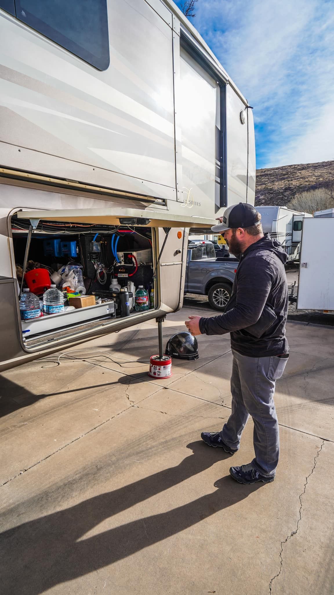 Man near a large RV with open compartment, reaching for something outdoors on a sunny day.