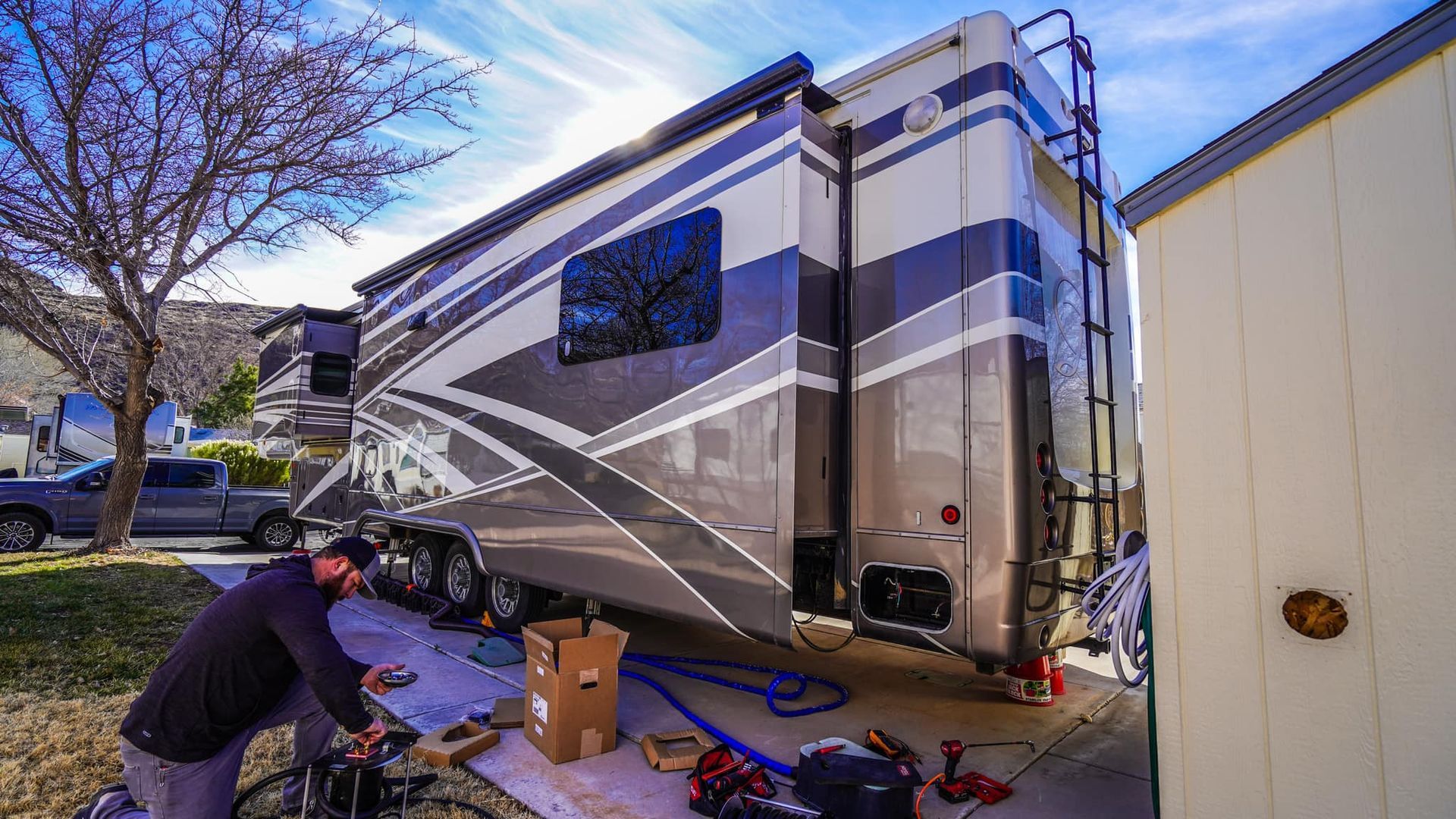 A man works on an RV parked on a driveway. Sun shines on the large, two-toned vehicle.