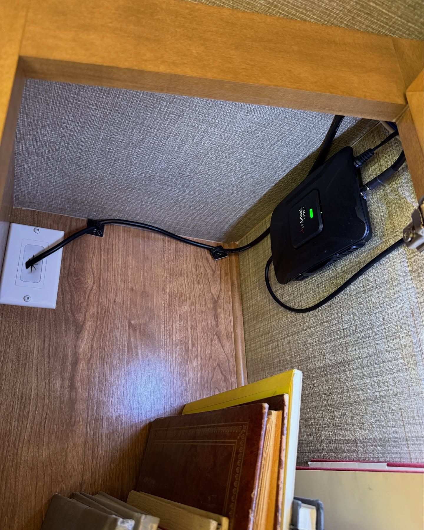 A black device with cords is mounted under a wooden shelf, next to a power outlet and books.