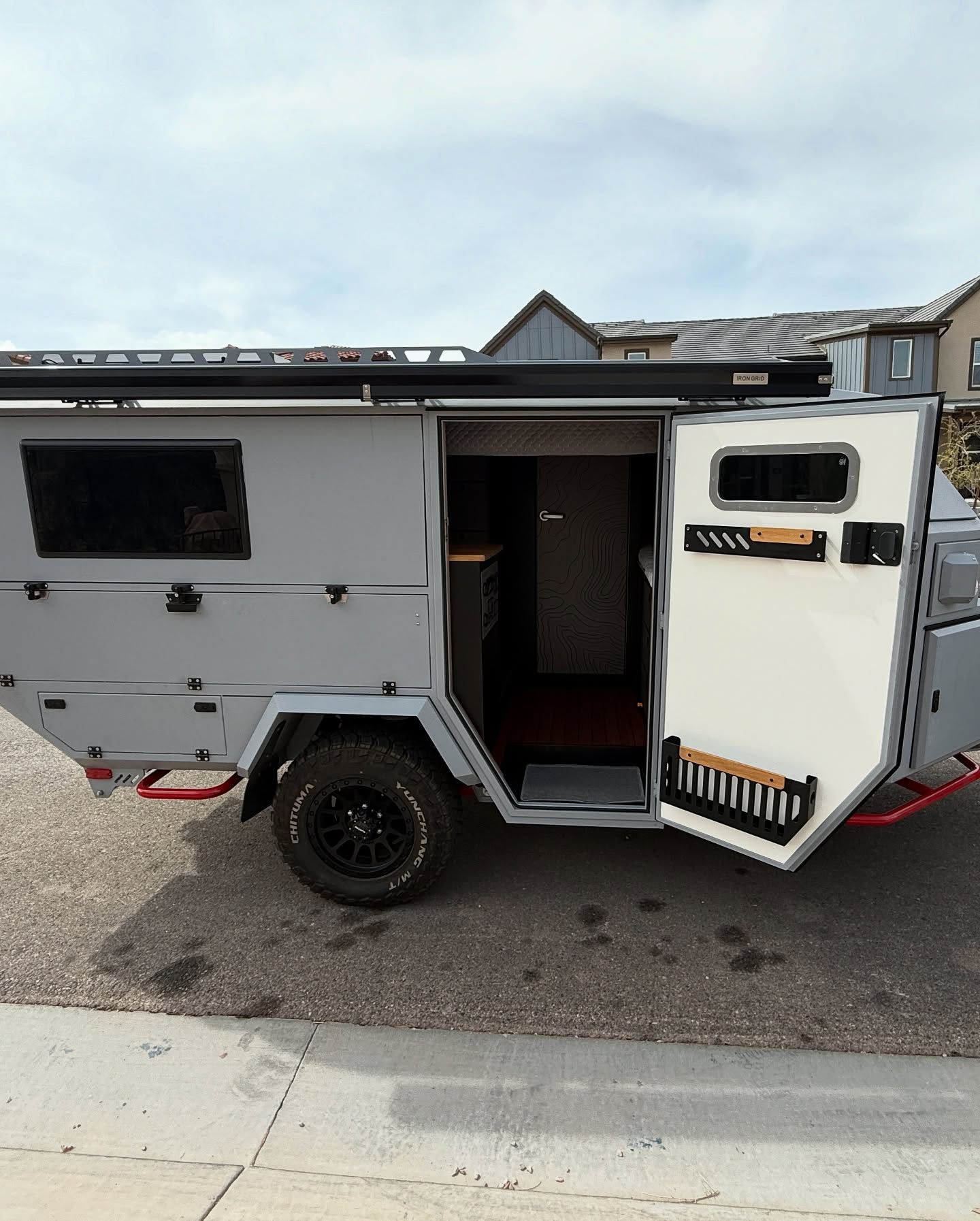 Gray and white off-road camper trailer parked on a street with its door open, showcasing its interior.
