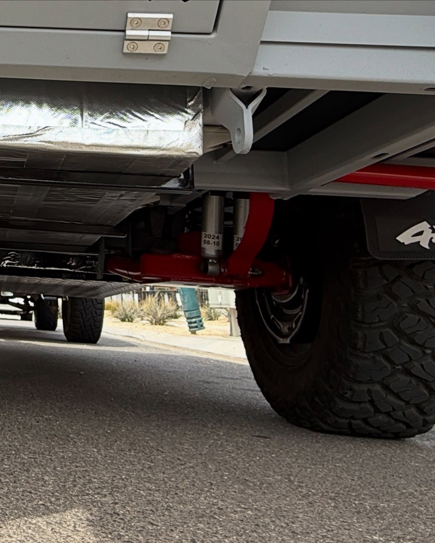 Undercarriage of an off-road trailer. Red suspension arms, shock absorbers, and large tire on asphalt.