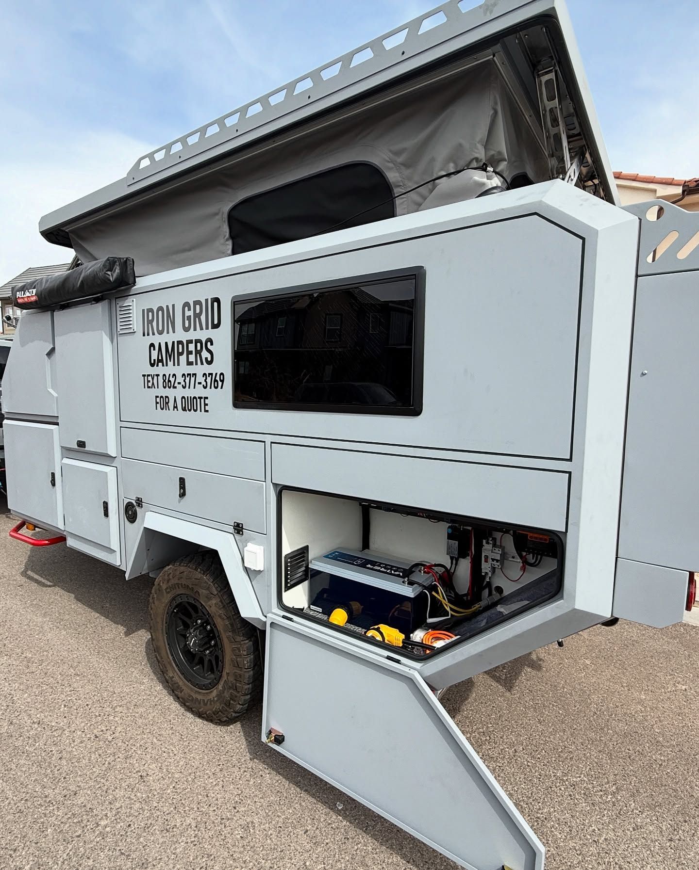 Gray off-road camper trailer with pop-up roof and open storage door, black wheels, against a daytime sky.