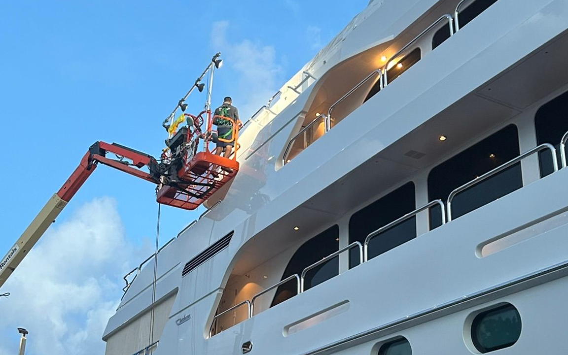 Workers on a lift washing the side of a large white yacht against a blue sky.