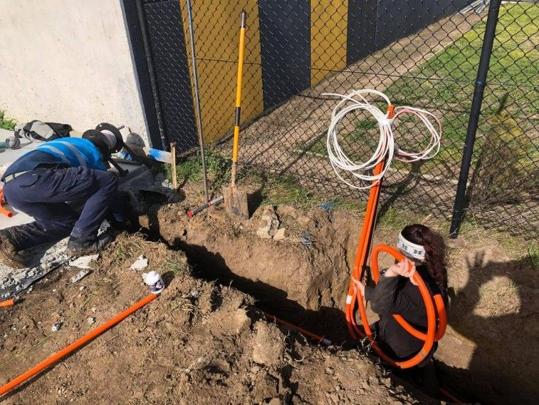 Two Workers Installing Orange Cables in a Trench Near a Fence.