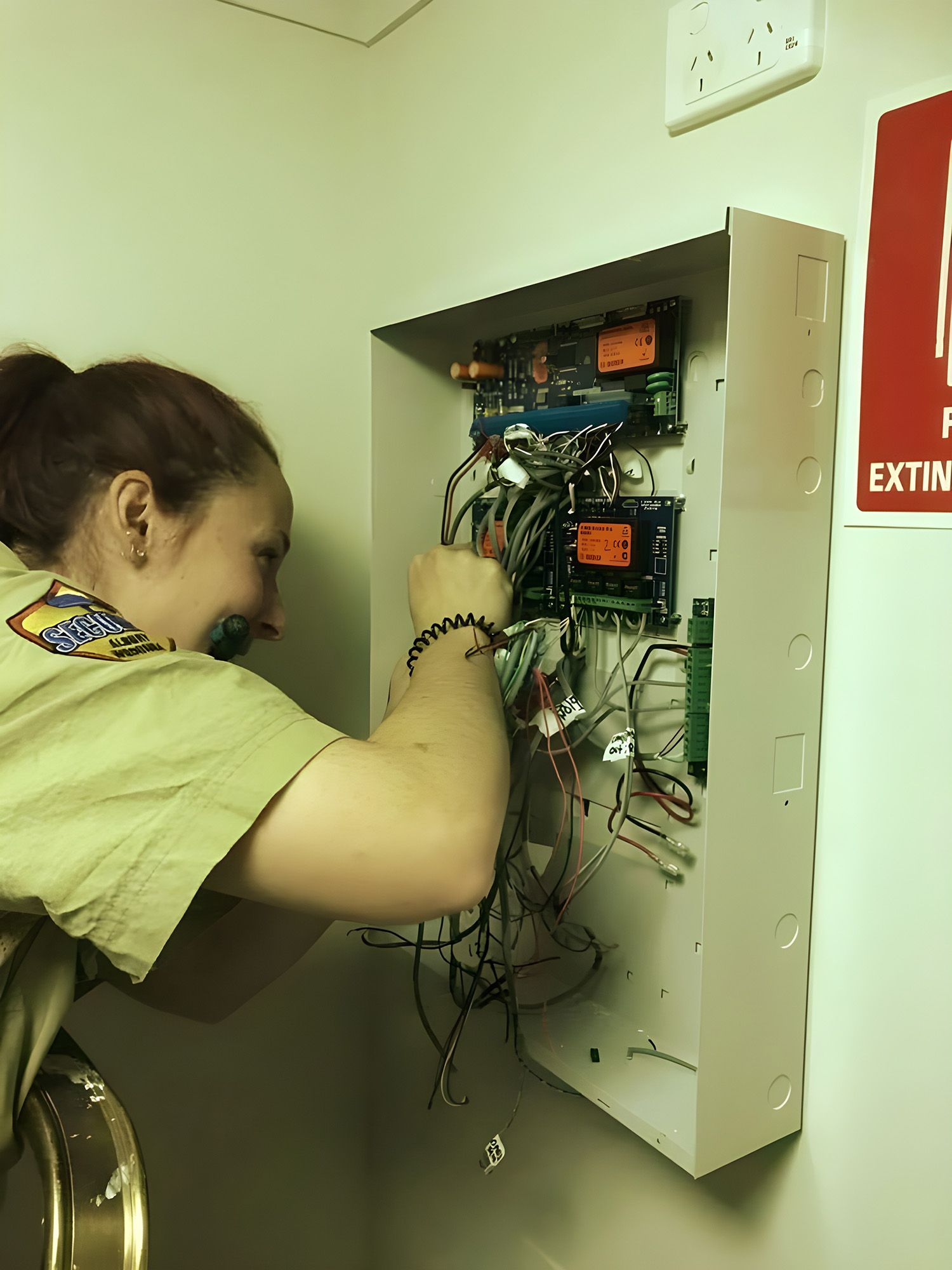 A Technician In A Tan Uniform Works On An Open Wall-Mounted Fire Alarm Control Panel