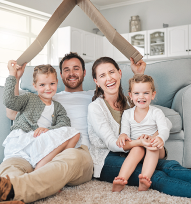 Family of four under a cardboard roof, smiling on a living room floor.
