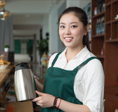 Woman in green apron holding a silver carafe, smiling in a cafe.