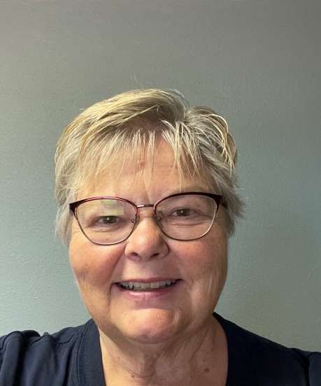 Woman with short blonde hair and glasses, smiling, wearing a dark top, neutral background.