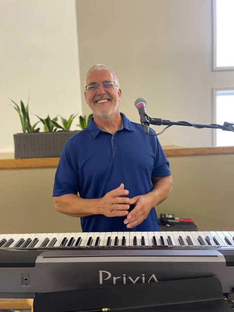 Man with gray hair, blue shirt, smiles at a piano with microphone in light-filled room.