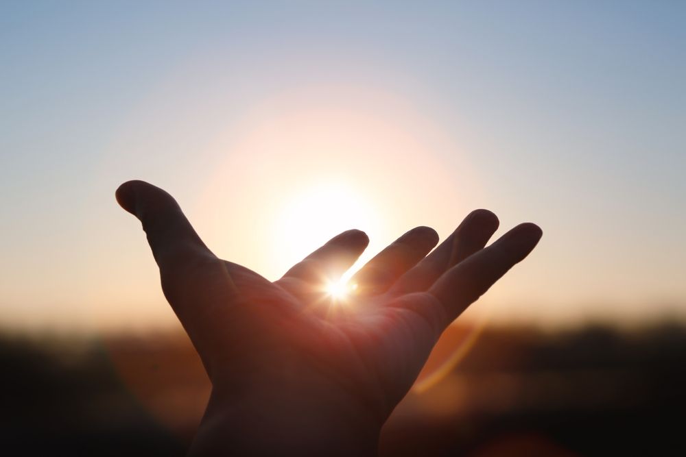 Hand reaching toward the sun, silhouetted against a warm, orange and blue sky.
