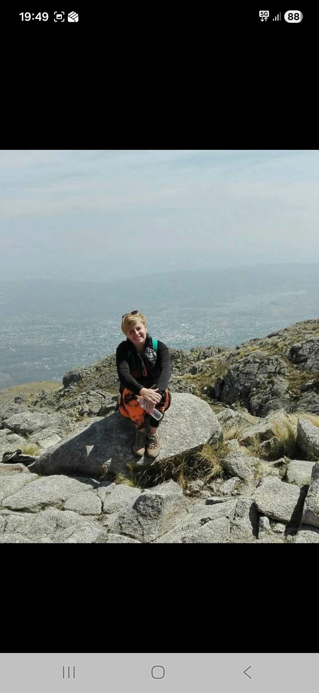 Una persona sentada en una roca en una montaña con vistas a una ciudad, sonriendo. Luz del día brillante.