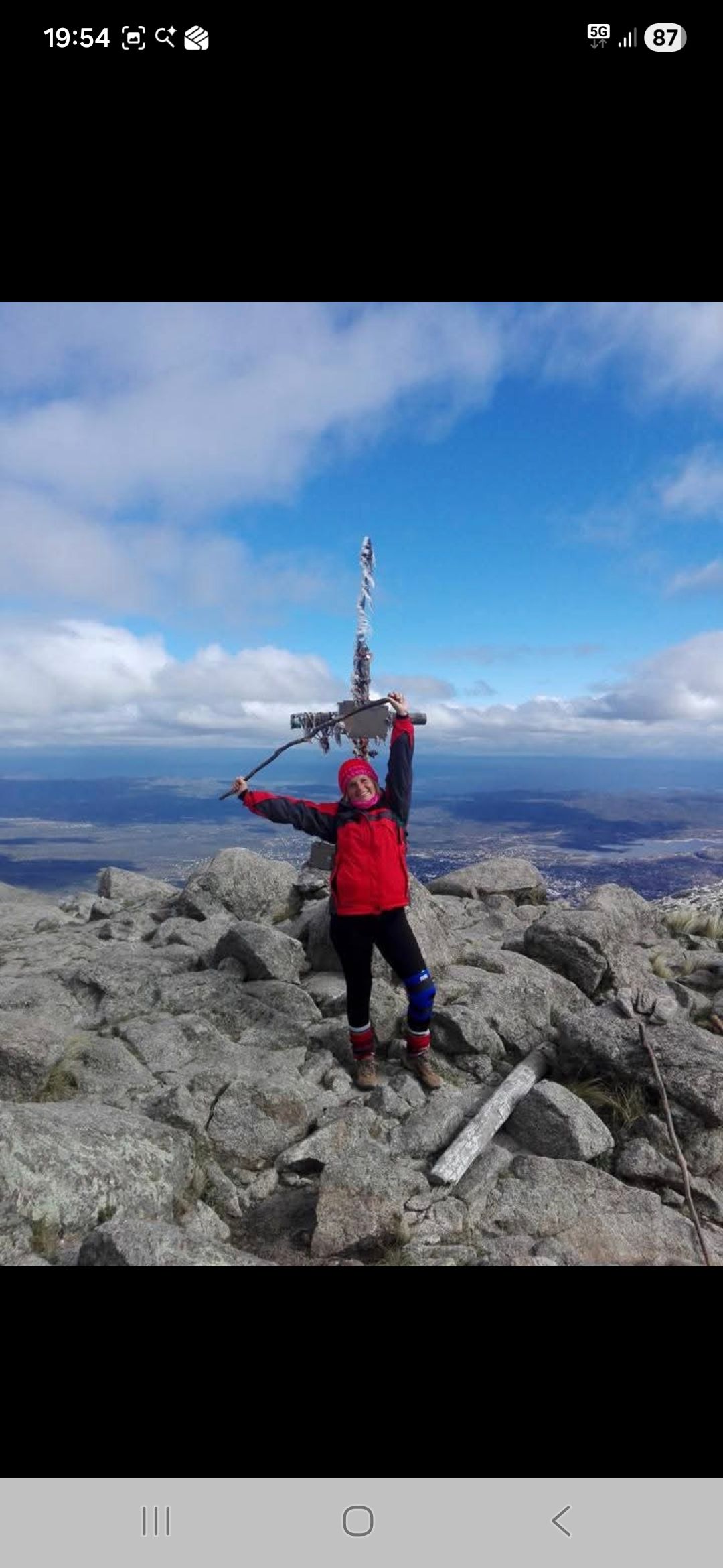 Persona con chaqueta roja en la cima de una montaña rocosa, levantando los brazos cerca de una torre, con cielo azul y nubes.