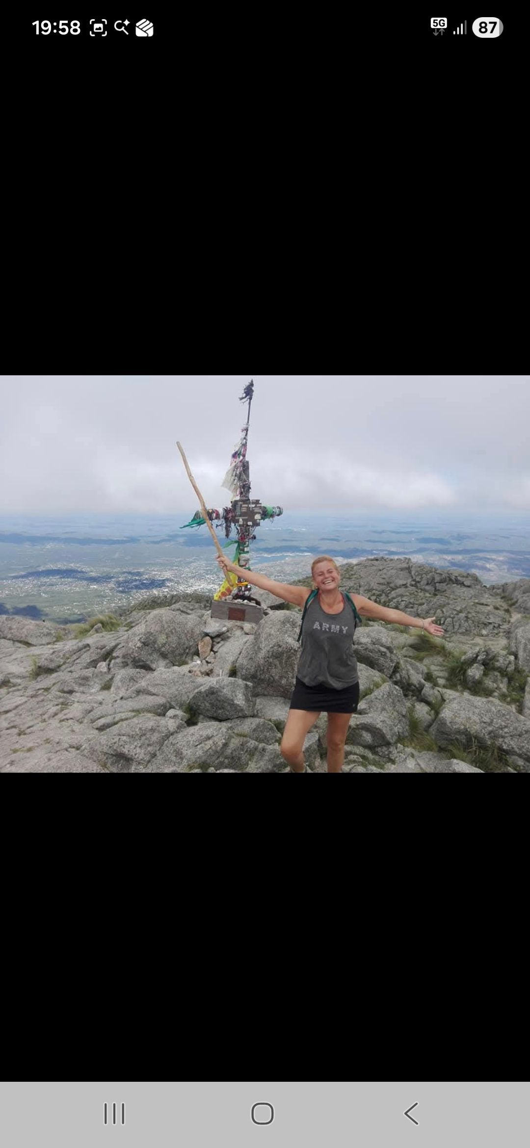 Mujer con los brazos extendidos en la cima de una montaña rocosa con una cruz y cielo nublado.