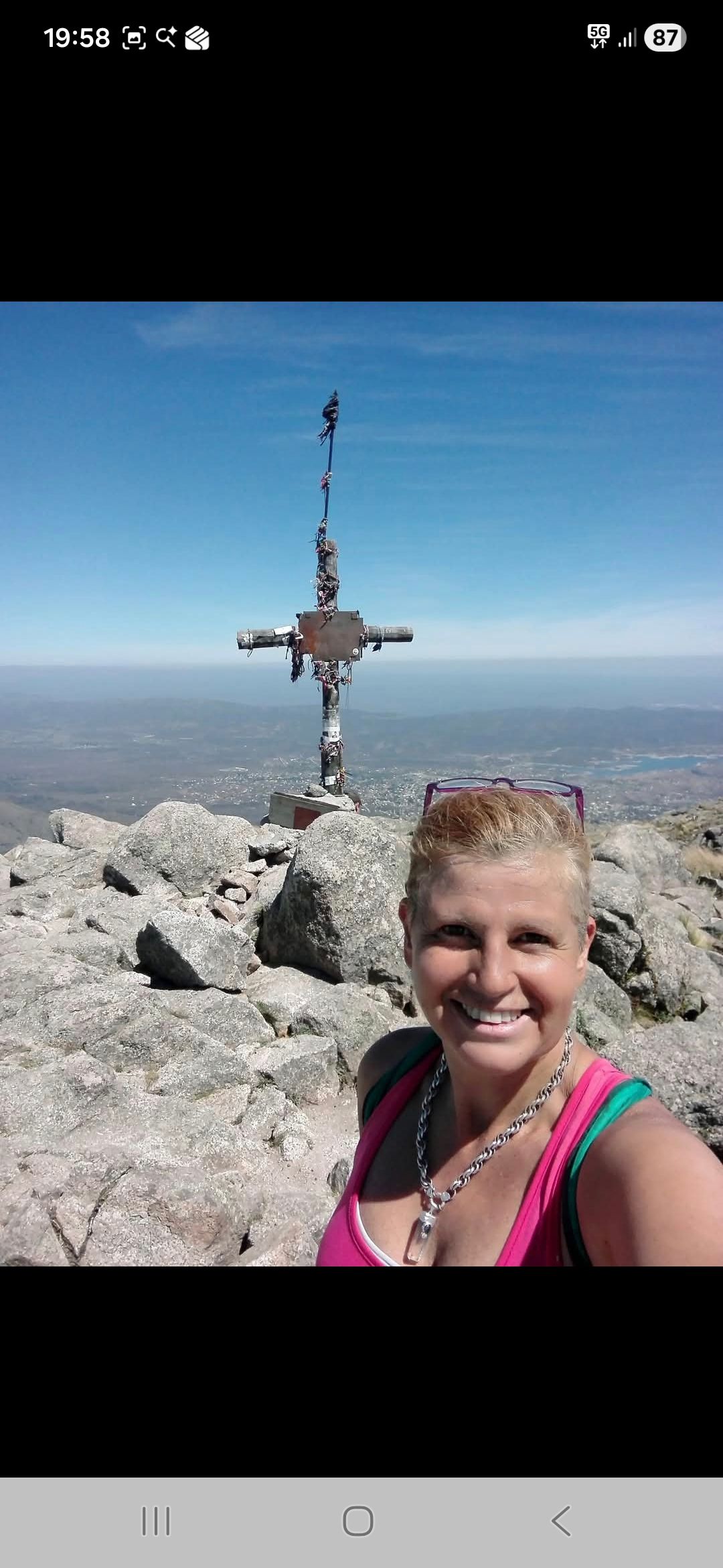 Una mujer sonríe a la cámara en la cima de una montaña rocosa con una cruz y un cielo azul claro.