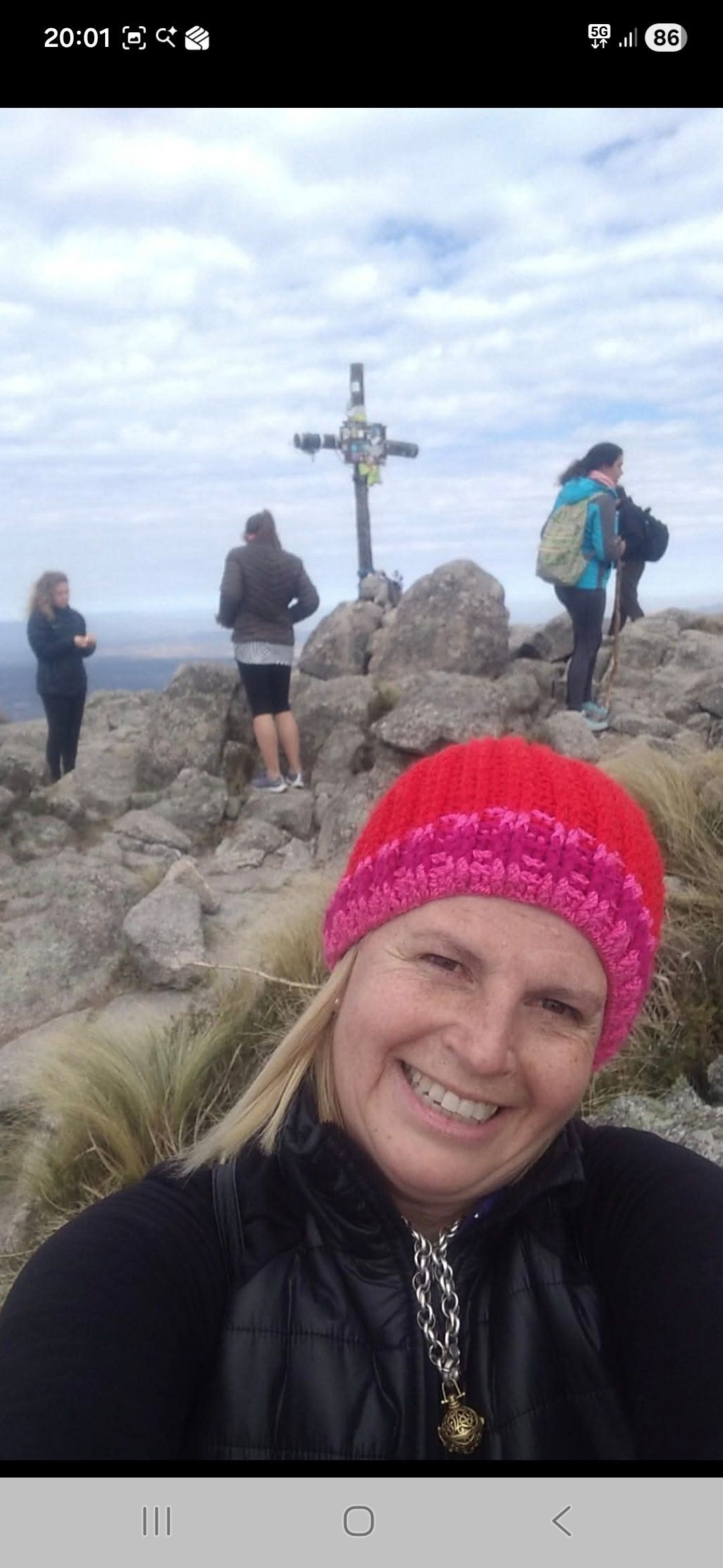 Una mujer con un sombrero rosa sonríe mientras se toma una selfi. Hay gente cerca de una cruz en la cima de una montaña.