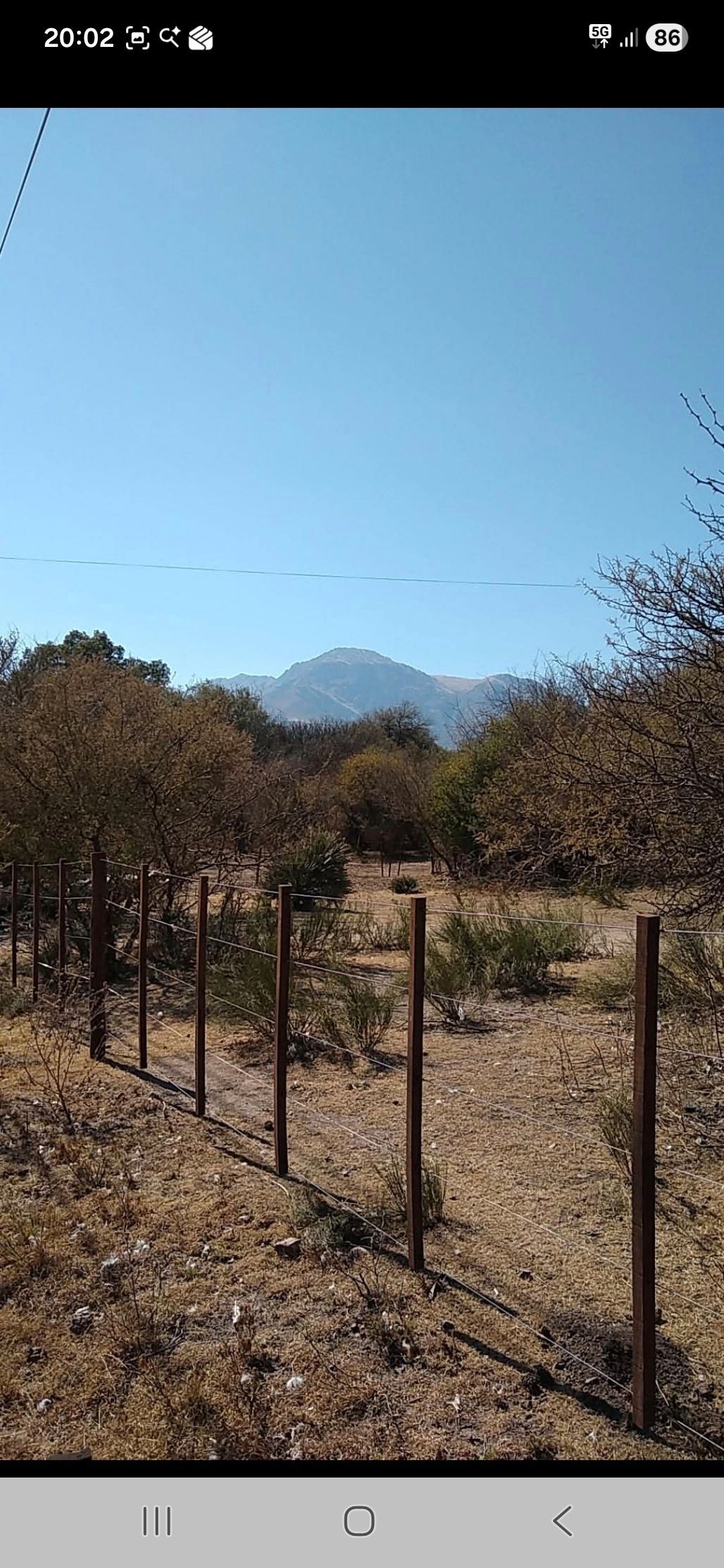 Un paisaje con una valla, árboles y una montaña bajo un cielo azul claro.