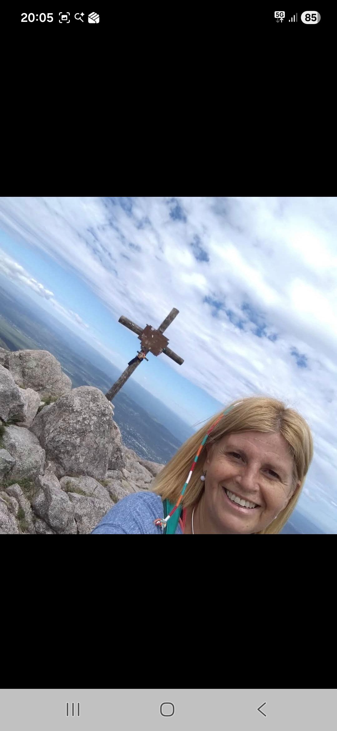 Una mujer sonríe a la cámara en la cima de una montaña con una cruz. Cielo azul con nubes de fondo.